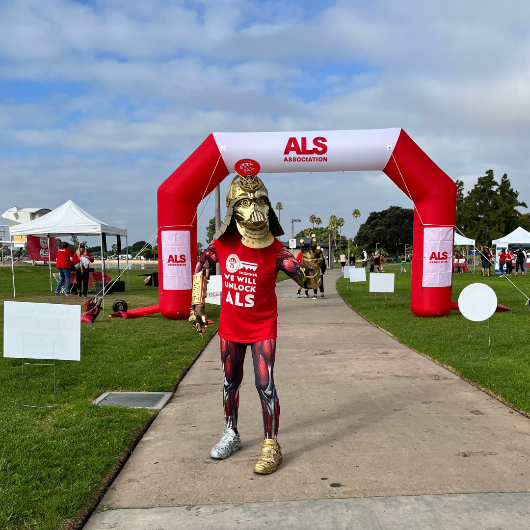 Person dressed in a Spartan warrior costume wearing a golden helmet, gold armor on arms, and athletic leggings, standing on a pathway at an ALS fundraising event with a large red and white ALS awareness arch and tents in the background.