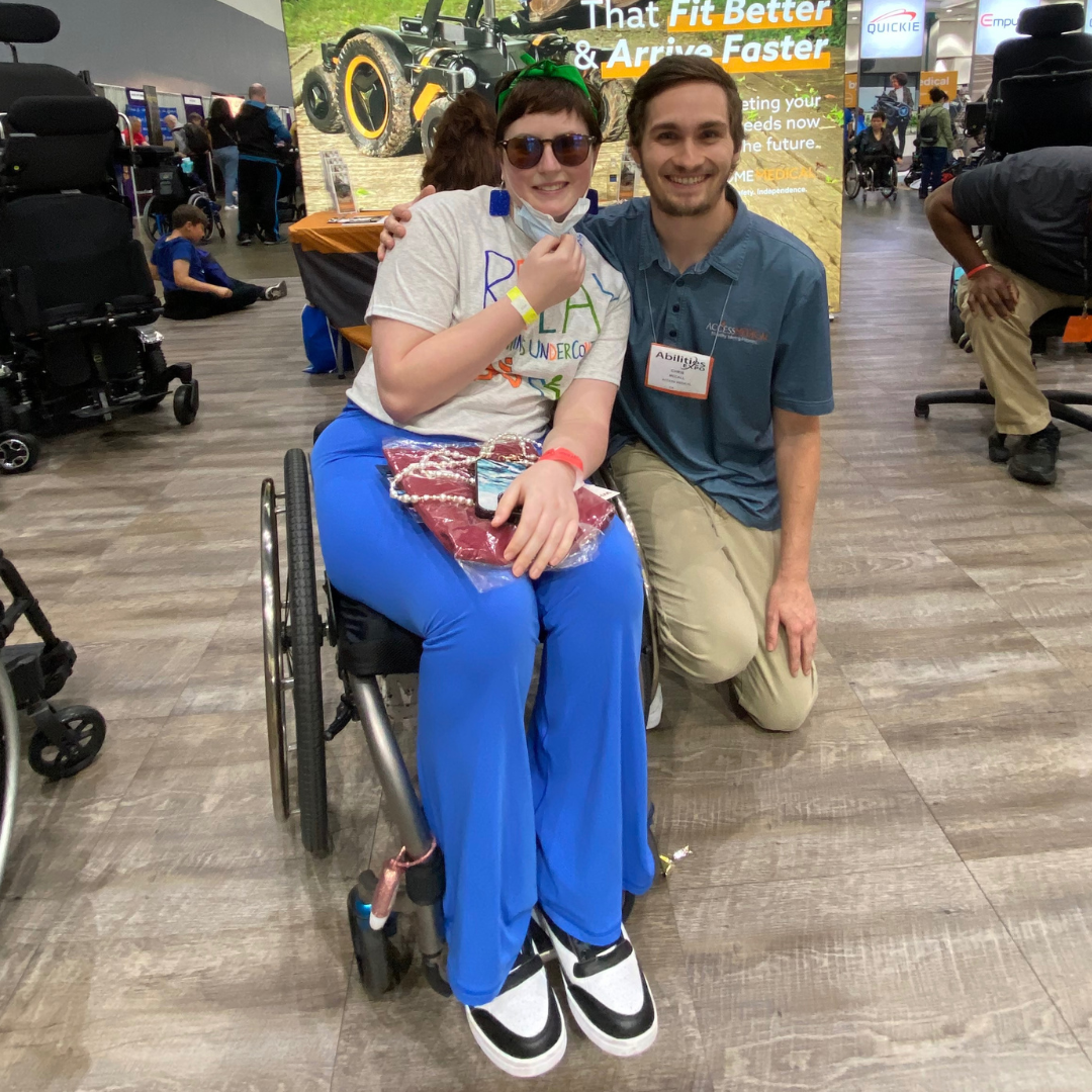 A young woman in a wheelchair and a man kneeling next to her smile for the camera in a busy indoor event space. The woman has short brown hair, sunglasses, and is wearing a white t-shirt with colorful writing, blue pants, and white sneakers. She is h
