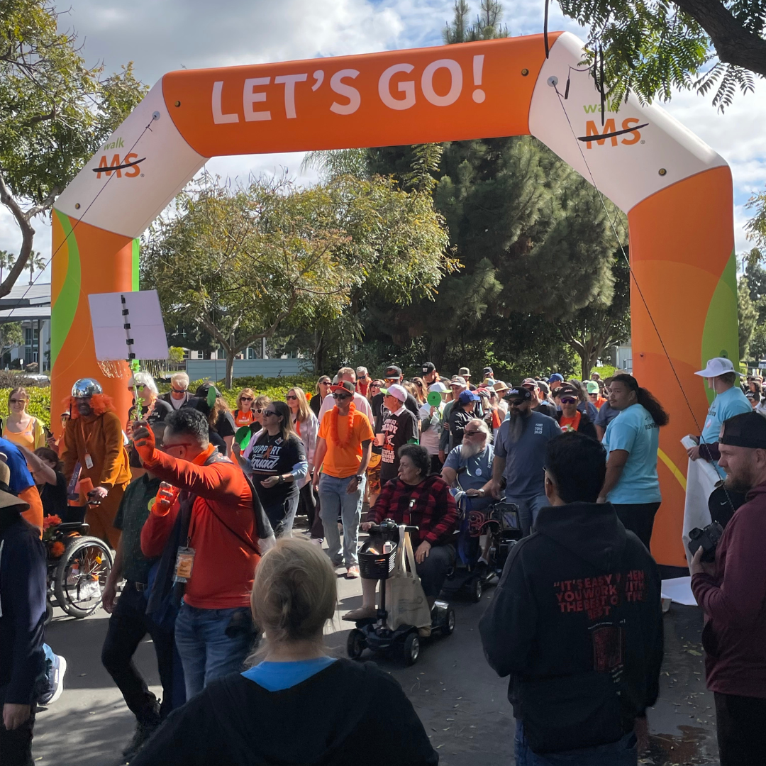 Crowd of people participating in a walk event under an orange and white archway with 'LET'S GO!' written on it, trees in the background, and partly cloudy sky.