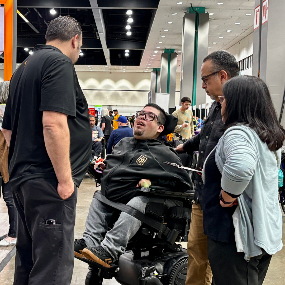 A group of four people having a discussion at a crowded indoor event. One person is seated in a motorized wheelchair, wearing glasses and a black jacket, looking up and speaking. The other three are standing around, listening and engaging in conversa