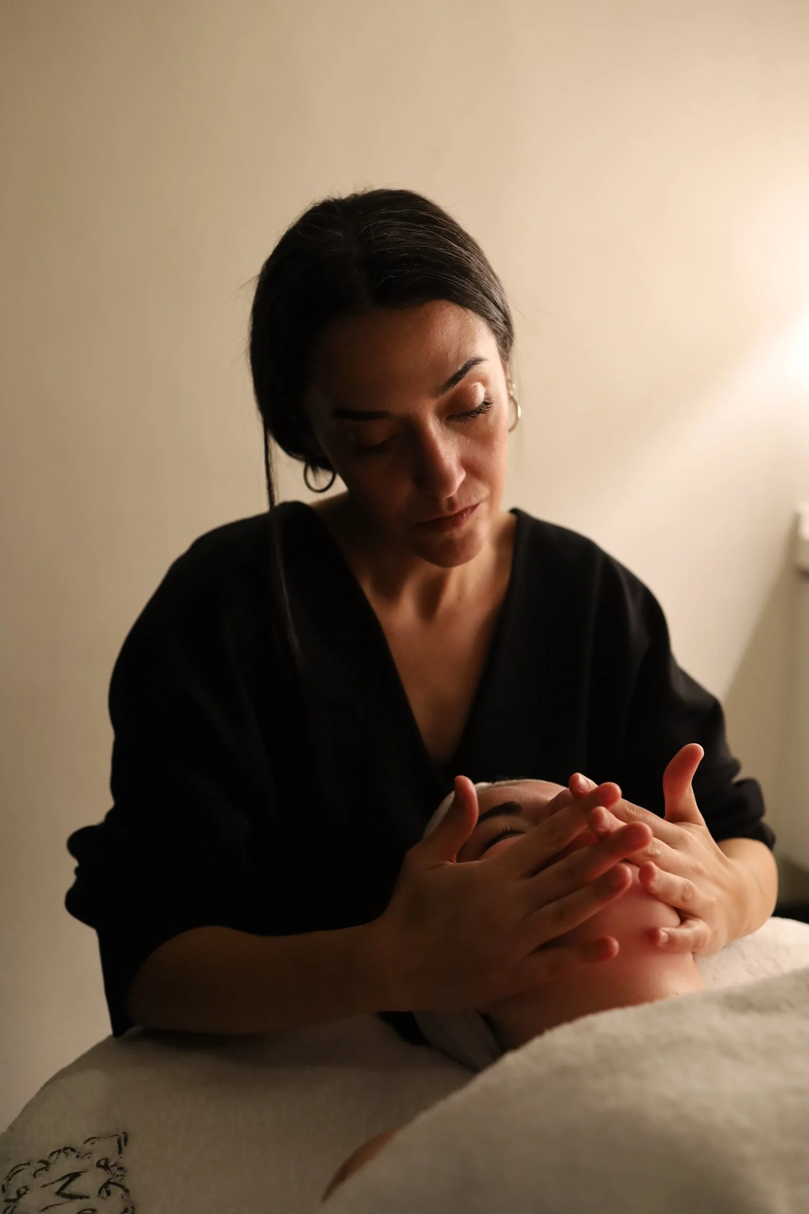 Una mujer con cabello largo y oscuro, peinada en ponytail, muestra productos de cuidado de la piel sobre su mano mientras sonríe en un ambiente de interior con cortinas beige.