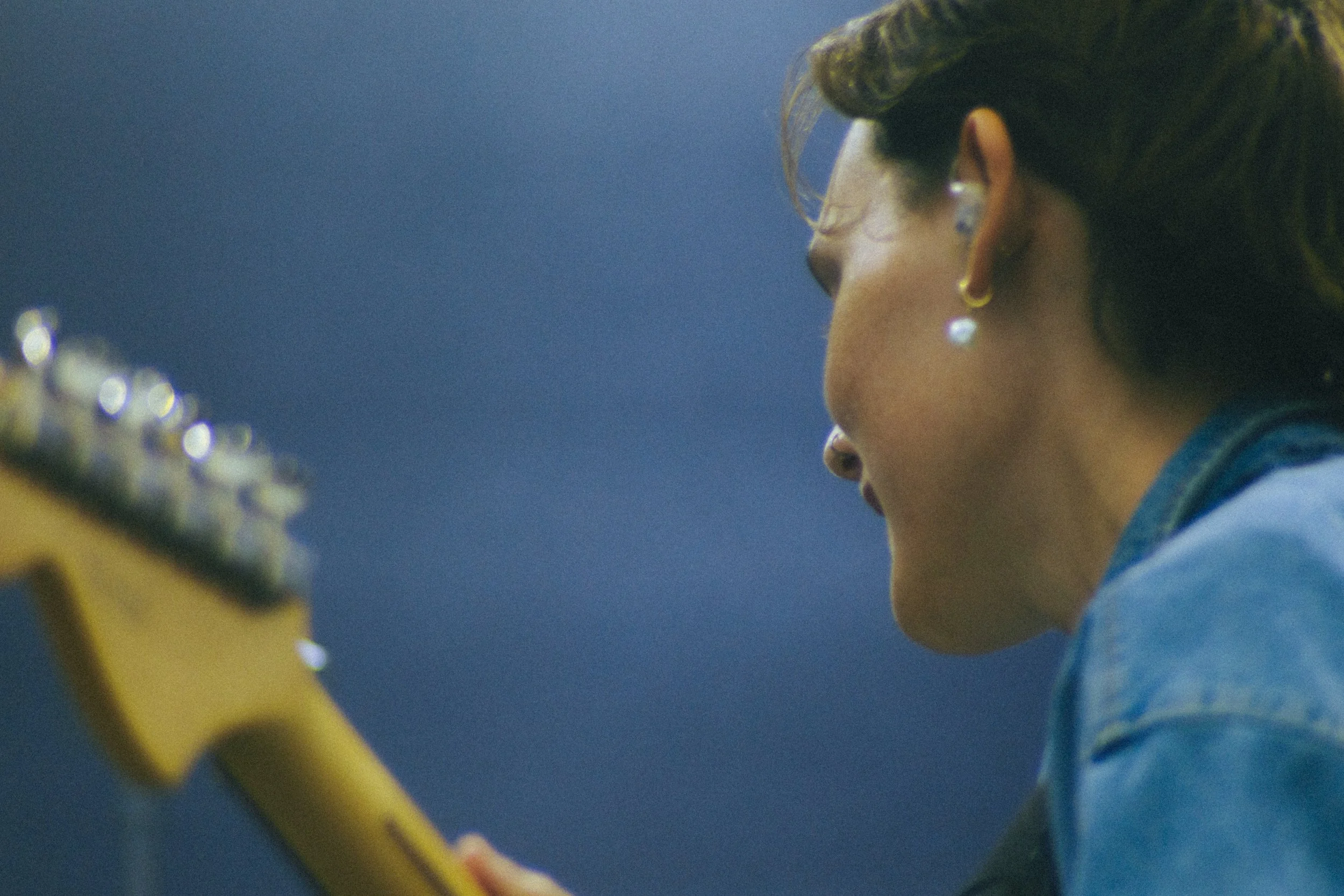 A person with short hair wearing earrings and a denim jacket, playing an electric guitar.