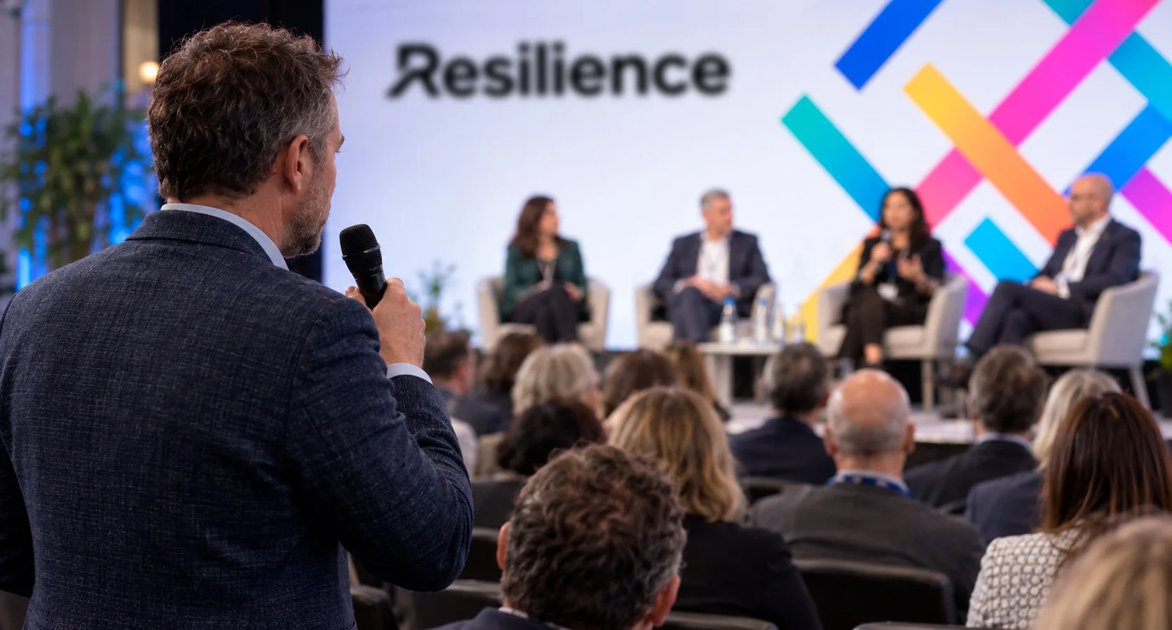 A man in a suit holding a microphone asks a question at a conference with a panel of four speakers on stage, behind which a large screen displays the word 'Resilience' and a colorful geometric pattern.
