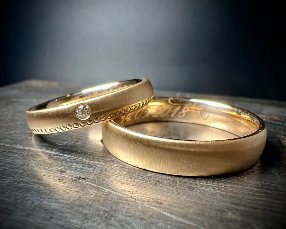 Zwei goldene Eheringe, einer mit Diamant, auf einer Holzoberfläche, dunkler Hintergrund.