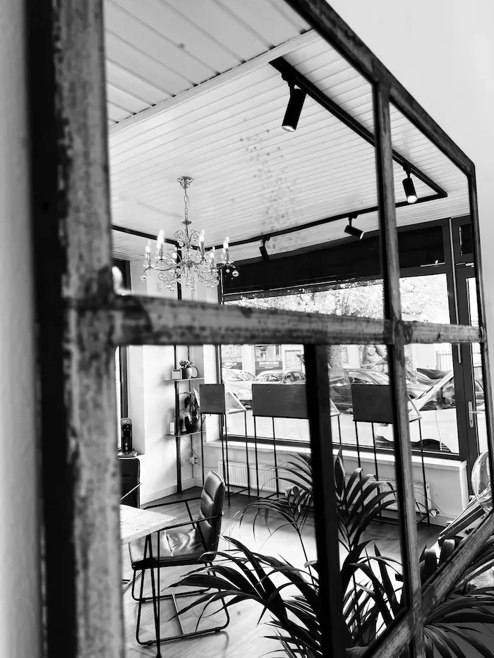 Ein Blick auf einen Raum durch ein Gitterfenster, mit einem Tisch, Stühlen, Pflanzen, einer Stehlampe und großen Fenstern, die den Blick auf parkende Autos nach draußen freigeben.