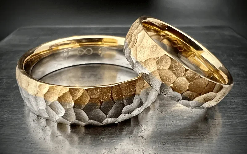 Zwei Eheringe mit einer gehämmerten Oberfläche, innen goldfarben, auf einem metallischen Untergrund