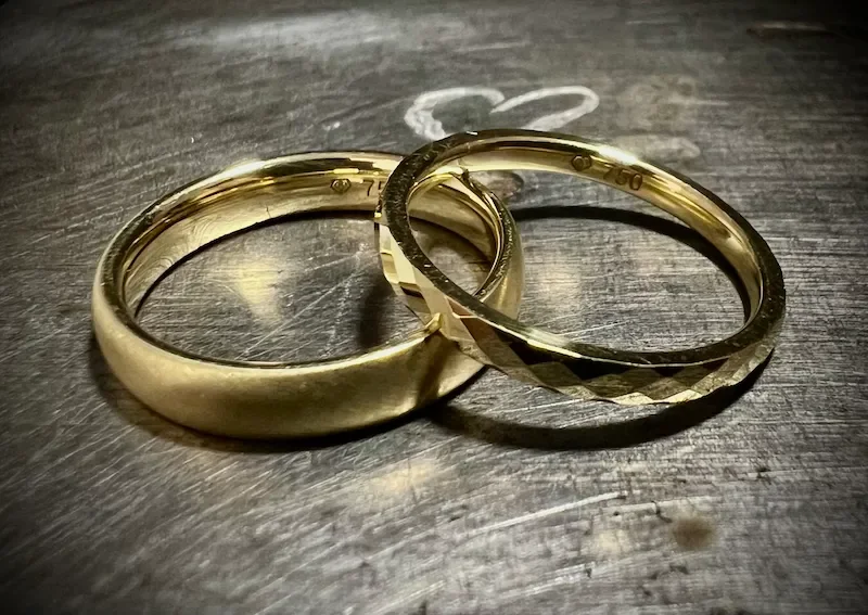 Zwei Goldringe liegen auf einer dunklen, metallischen Oberfläche.