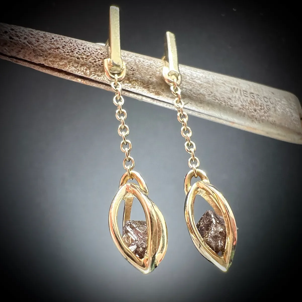 Paargoldohrringe aus recyceltem Gold mit Diamanten hängen an einer Metallstange, dunkler Hintergrund.