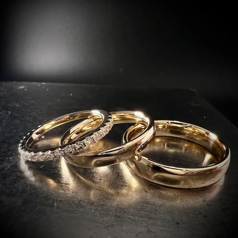 Drei goldene Ringe, einer mit Diamanten besetzt, auf einer schwarzen Oberfläche.