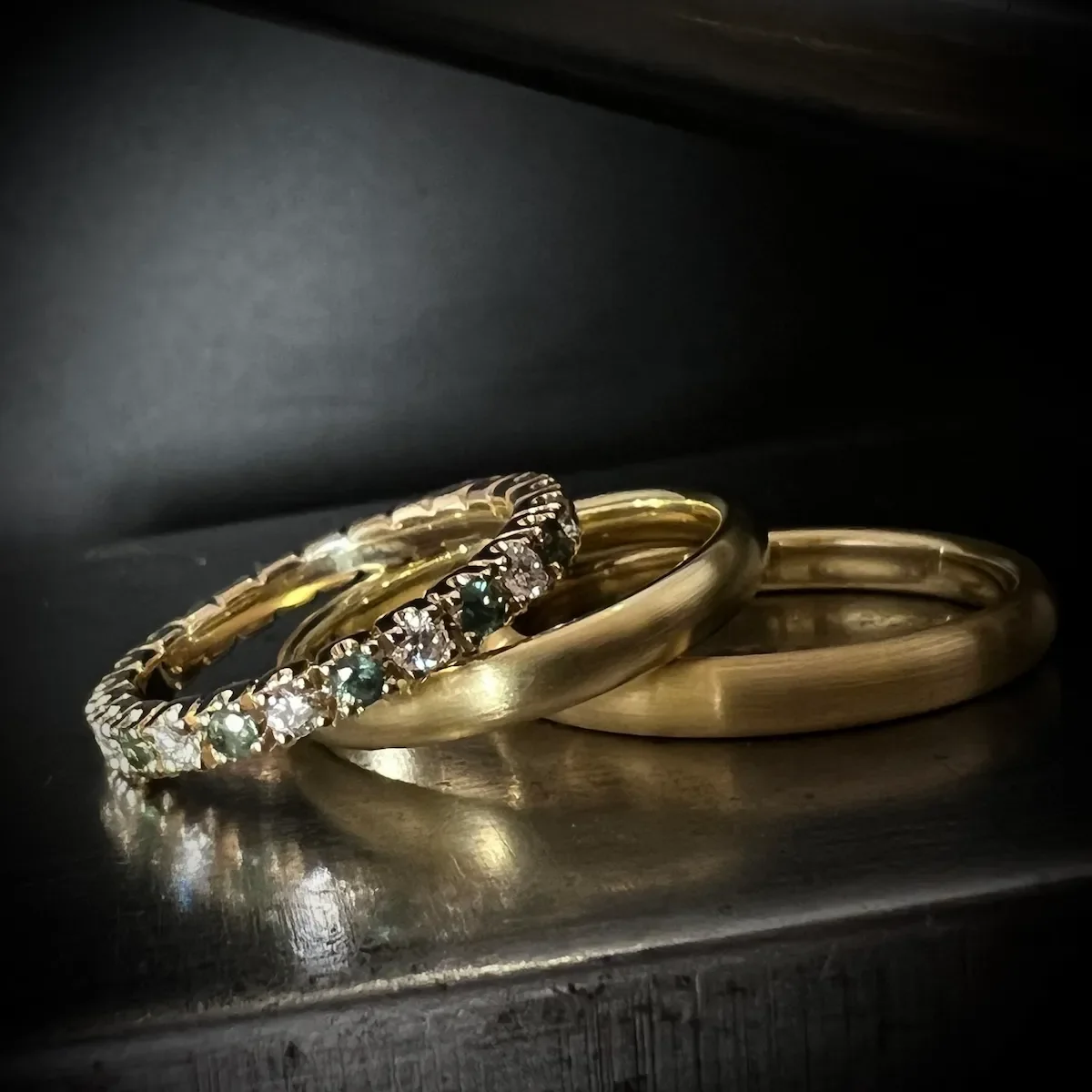 Vier goldene Ringe, eine mit Edelsteinen und Diamanten, liegen auf einer dunklen Oberfläche.