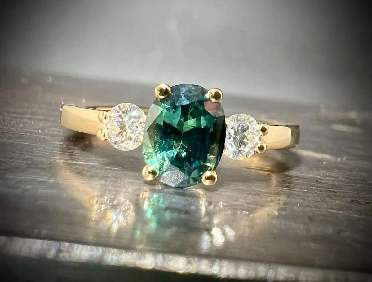  Eleganter Verlobungsring aus recyceltem Gelbgold mit einem ovalen, tiefgrünen Saphir und zwei seitlichen Diamanten im Brillantschliff