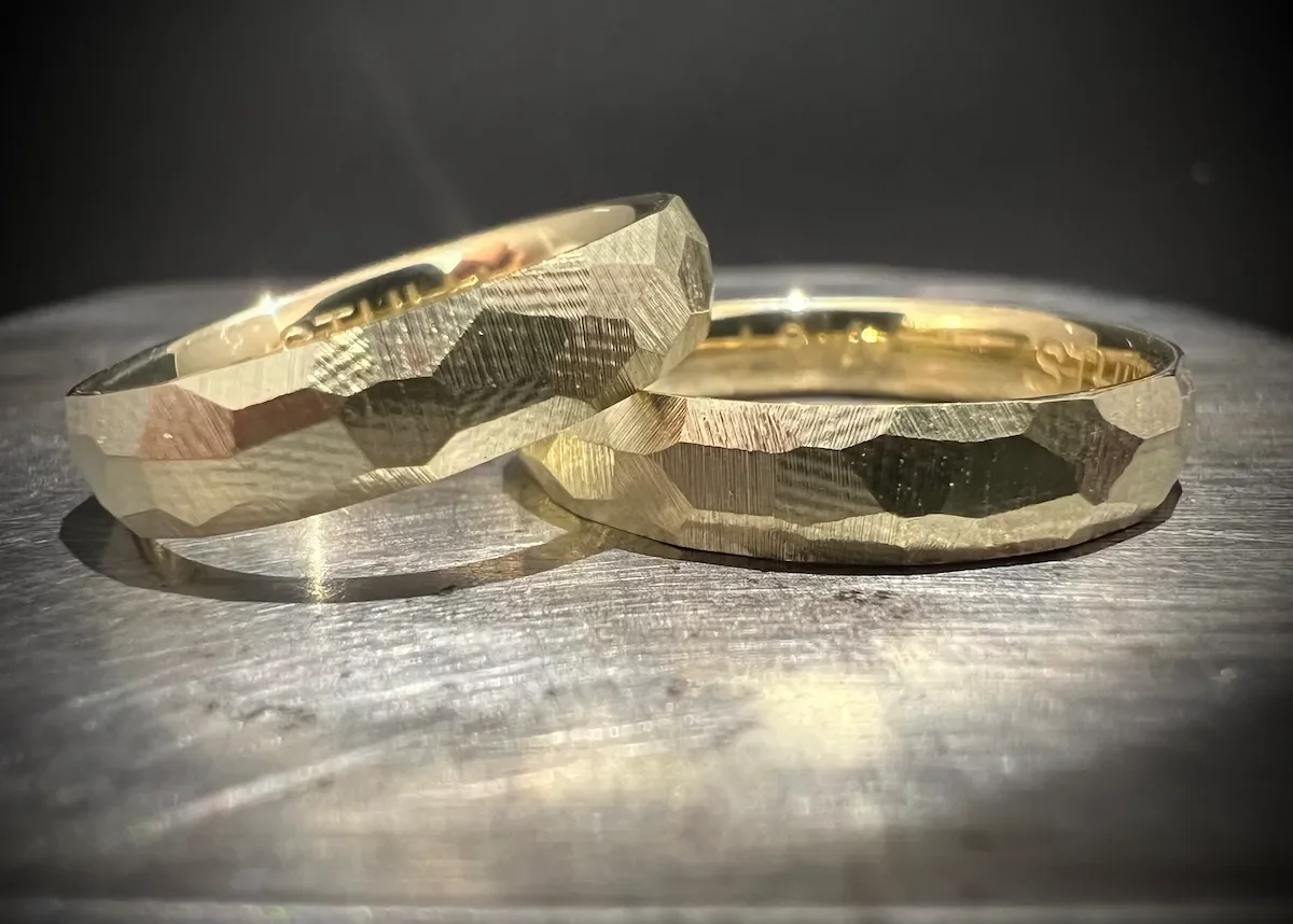 Zwei goldene Eheringe mit eingravierten Muster liegen auf einer metallic Oberfläche, beleuchtet von seitlichem Licht.