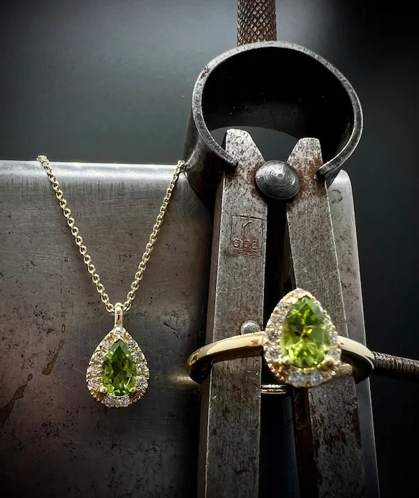 Ein Goldring und eine Halskette mit grünen Peridot, eine Zange und ein Metallblock als Hintergrund.