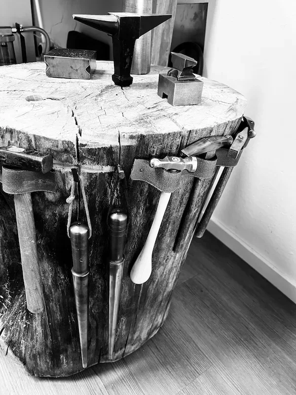 Schmiedewerkzeug auf einem Holzblock, darunter Hämmer und Werkzeuge hängen an der Seite.