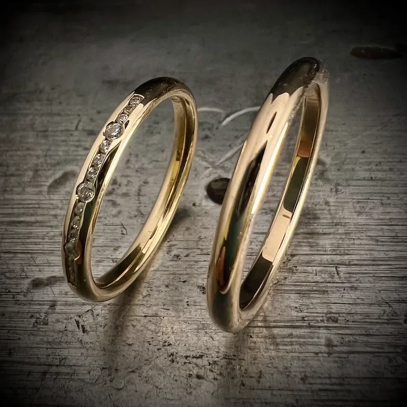 Zwei goldene Eheringe, einer mit kleinen Diamanten verziert, liegen auf einem Holzuntergrund.