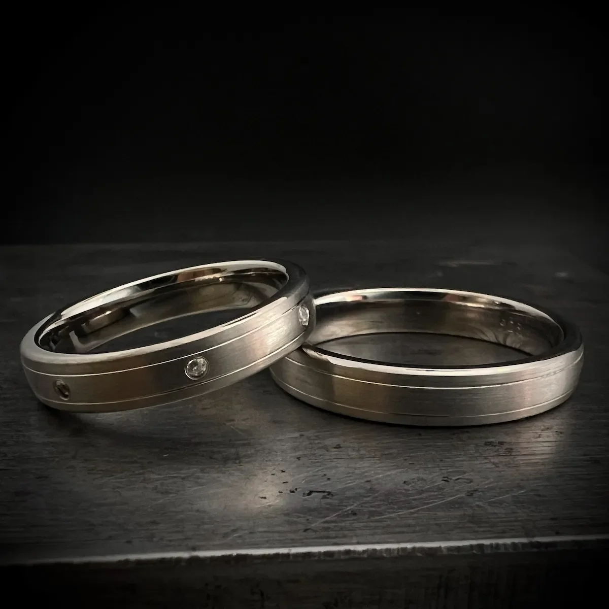 Zwei Platin Eheringe mit kleinen Diamanten auf einem dunklen Holztisch gegen schwarzen Hintergrund.
