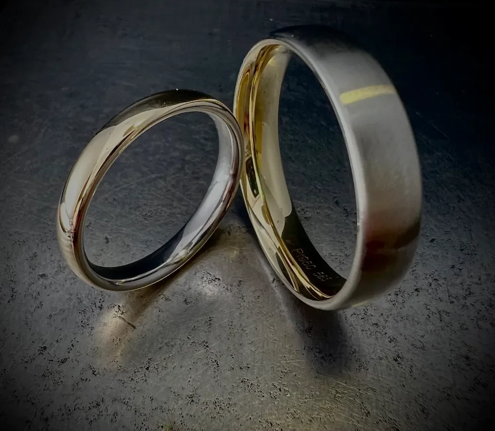 Zwei metallene Eheringe auf einer dunklen Oberfläche, leicht schräg positioniert.