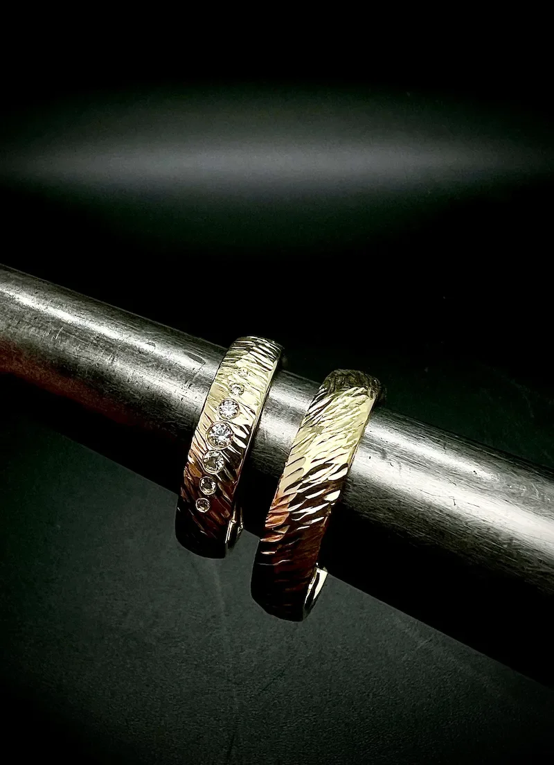 Zwei Silberschmuckringe mit unterschiedlichen Oberflächenstrukturen, einer mit kleinen Diamanten, die auf einem Metallrohr liegen.