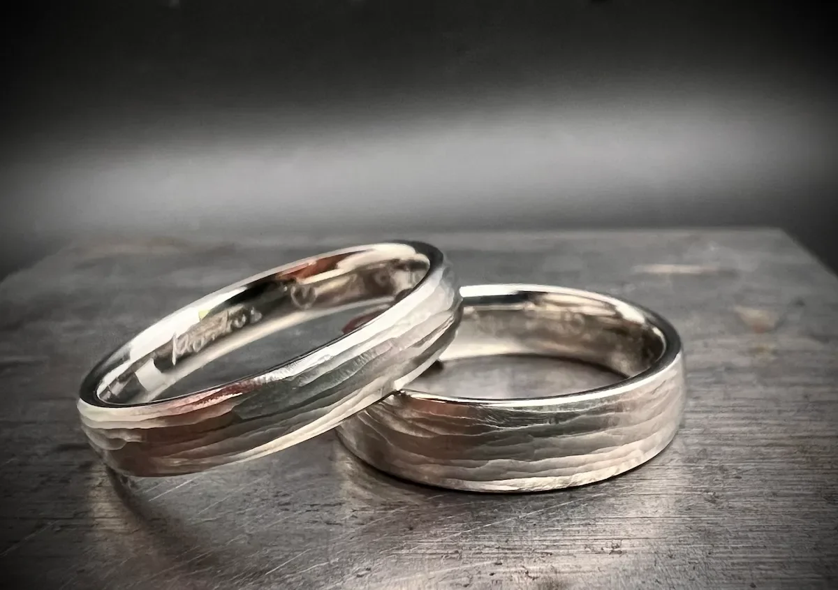 Zwei Eheringe aus rotgoldenem und silbernem Metall liegen auf einer Holztischplatte, leicht übereinander gelegt.