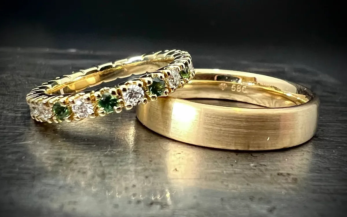 Zwei goldene Eheringe, einer mit Diamanten und grünen Edelsteinen, auf einer metallischen Oberfläche.