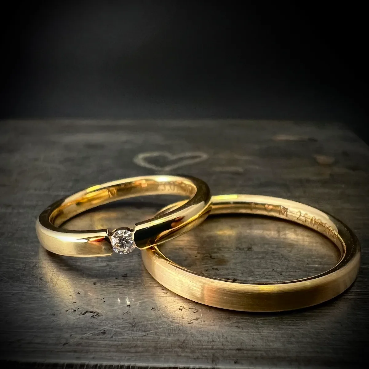 Zwei goldene Eheringe, einer mit Diamant, liegen auf einem Holzbrett, im Hintergrund ist eine kleine Herzzeichnung zu sehen.