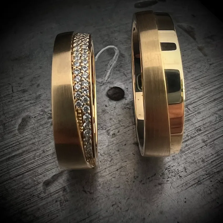 Zwei goldene Armreife liegen auf einem Holztisch, einer mit Steinen besetzt.