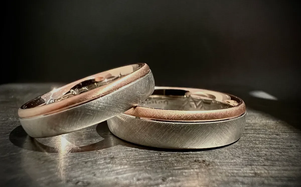 Zwei miteinander verbundene Eheringe aus Silber und Gold auf einer dunklen Holzoberfläche.