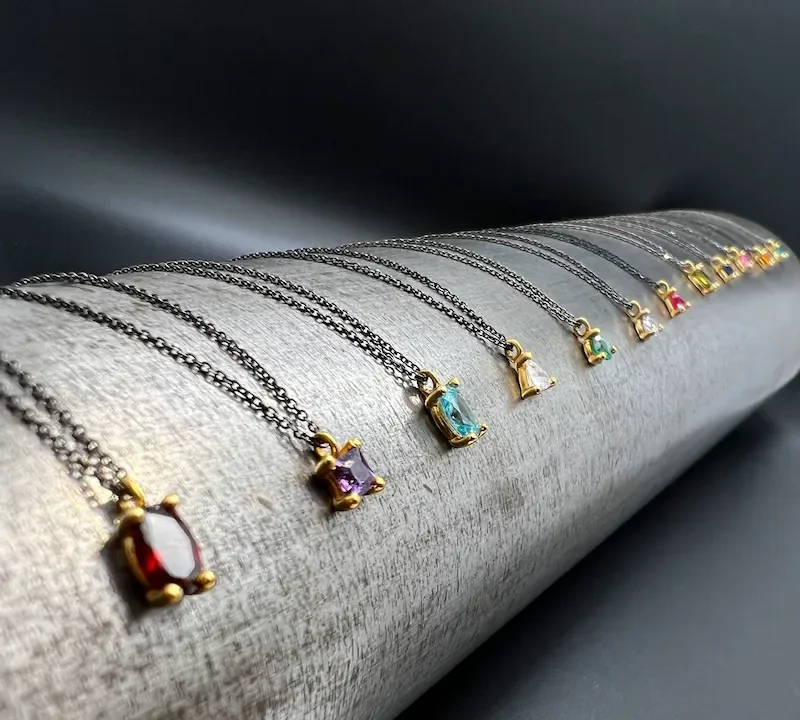 Eine Reihe von Halsketten mit kleinen, bunten Edelsteinanhängern in goldenen Halterungen, präsentiert auf einem grauen Stoffrollen in dunklem Hintergrund.