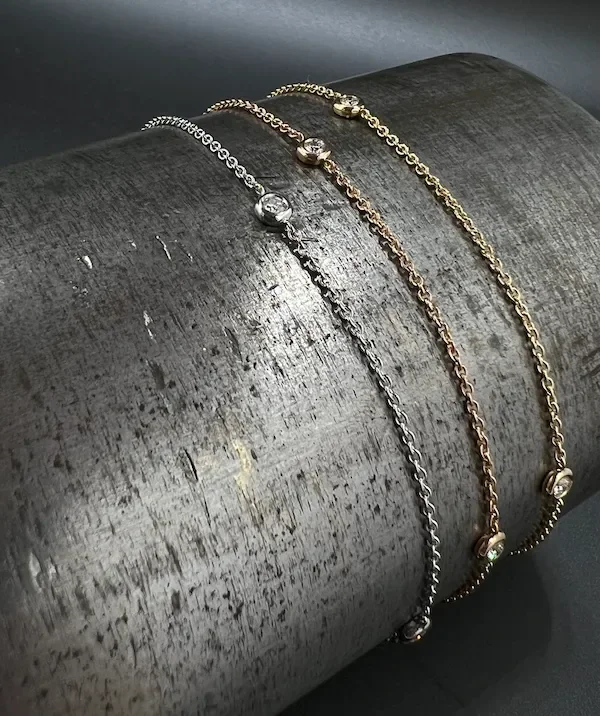 Drei silberne und goldene Armbänder mit kleinen Diamanten, auf einer grauen, glatten, runden Oberfläche gelegt.