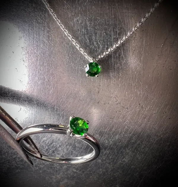 Silberne Halskette und Ring mit grünen Edelsteinen auf metallischer Hintergrundoberfläche.