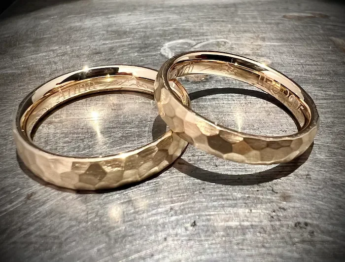 Zwei goldene Eheringe mit Hammer- oder Ringmuster auf einer dunklen, holzähnlichen Oberfläche.