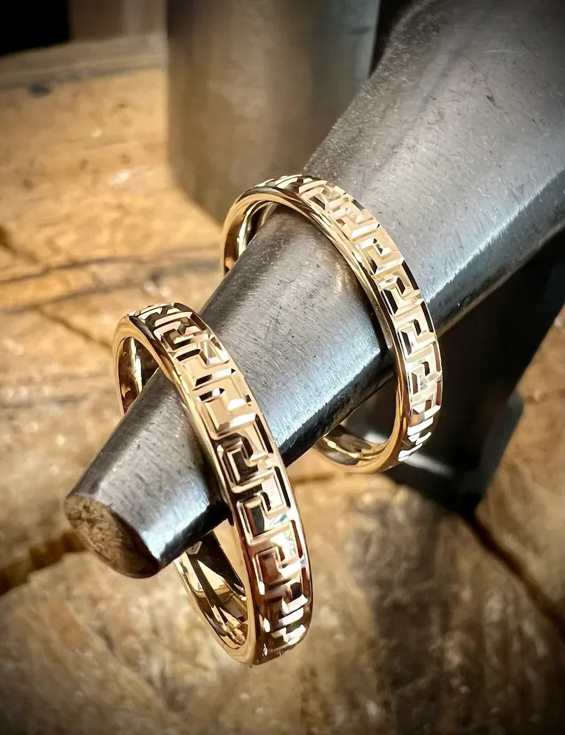 Zwei goldene Ringe mit einer geometrischen Musterung ruhen auf einem metallischen Gegenstand.