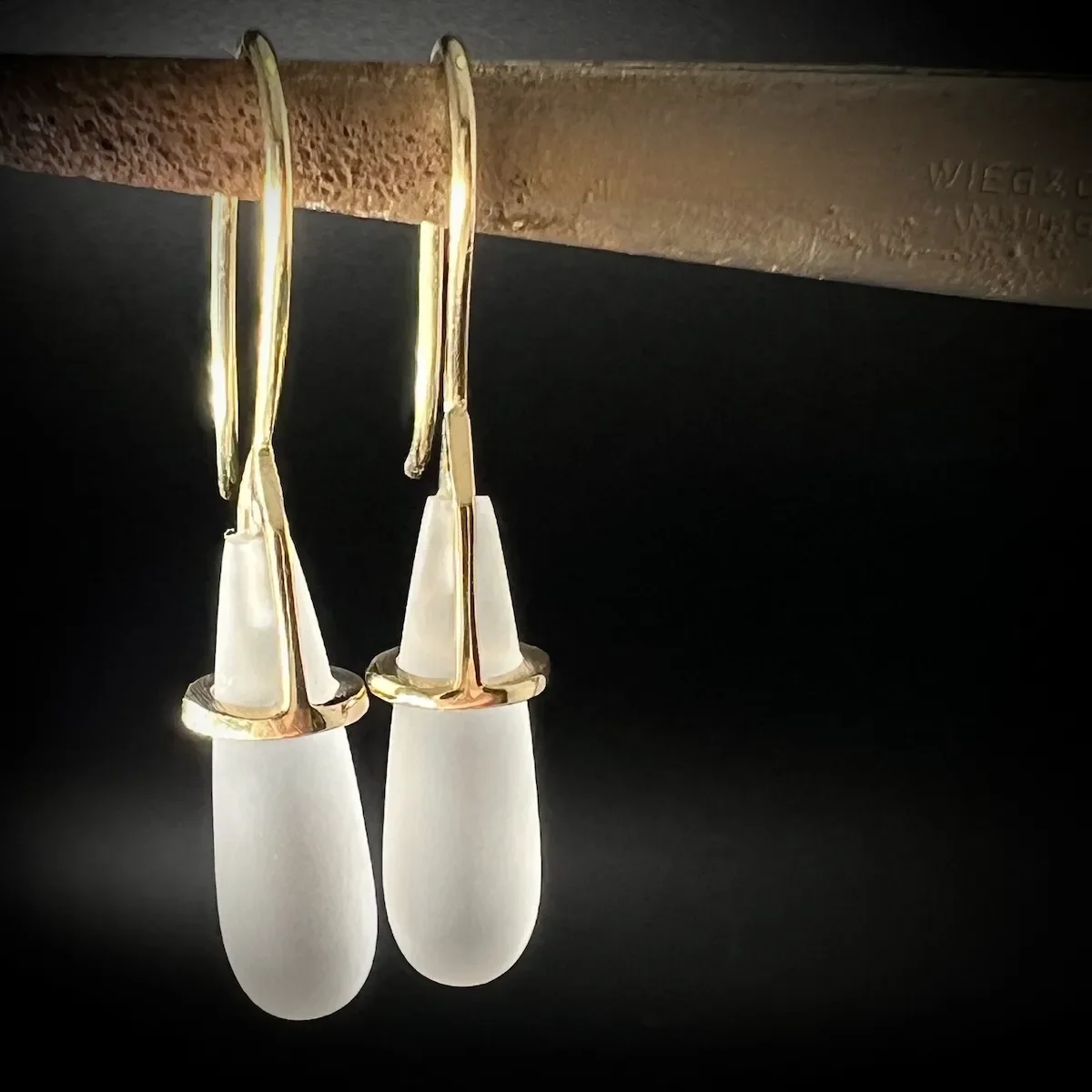 Zwei elegante Damen Ohrhänger mit weißen Tropfen-Design, hängen von einer Metallstange an einem schwarzen Hintergrund.