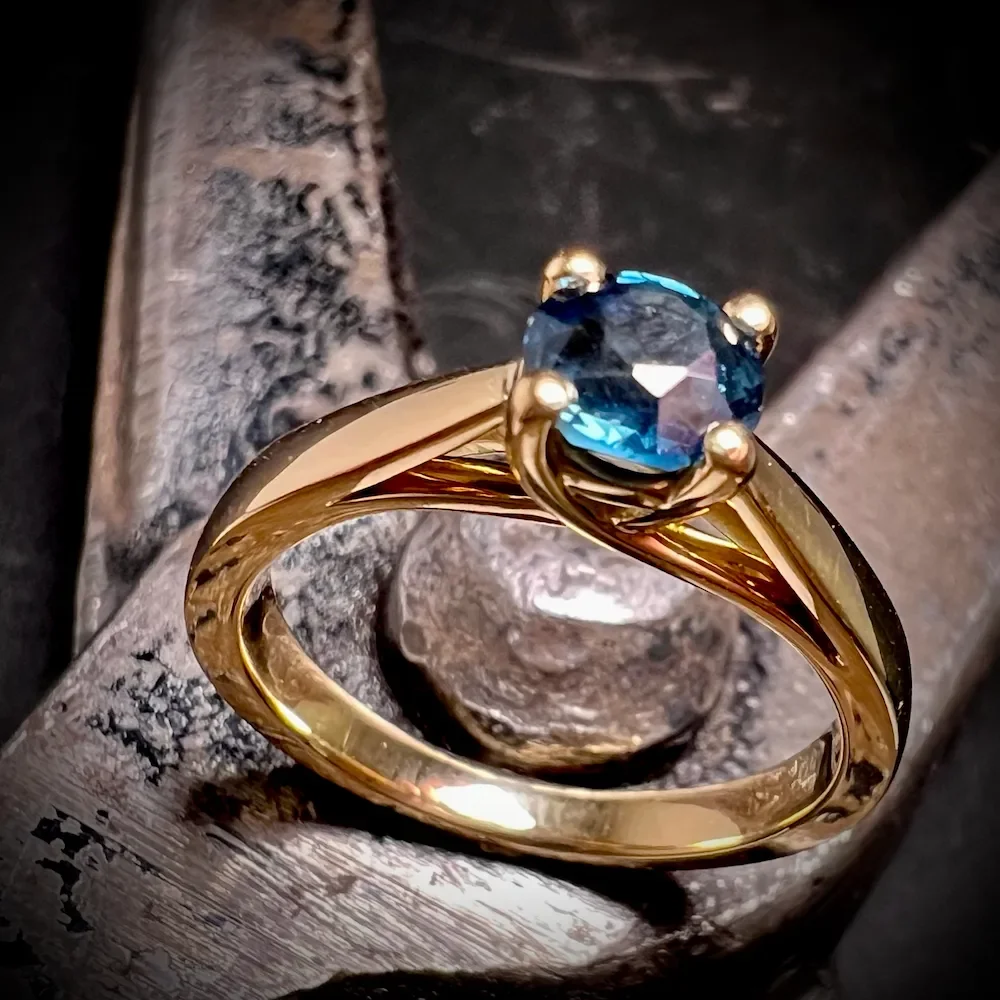 Gelbgold-Ring mit blauem Saphir in geschwungener Fassung auf Schmiedezange, handgefertigt in München.