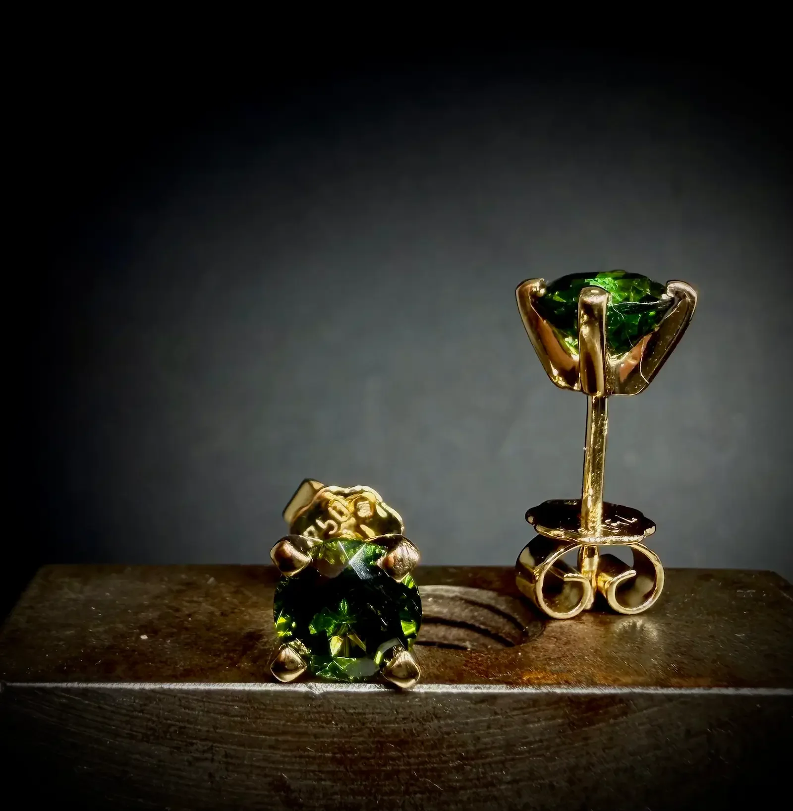 Zwei goldene Ohrringe mit grünen Saphir auf einer Holzoberfläche, dunkler Hintergrund.