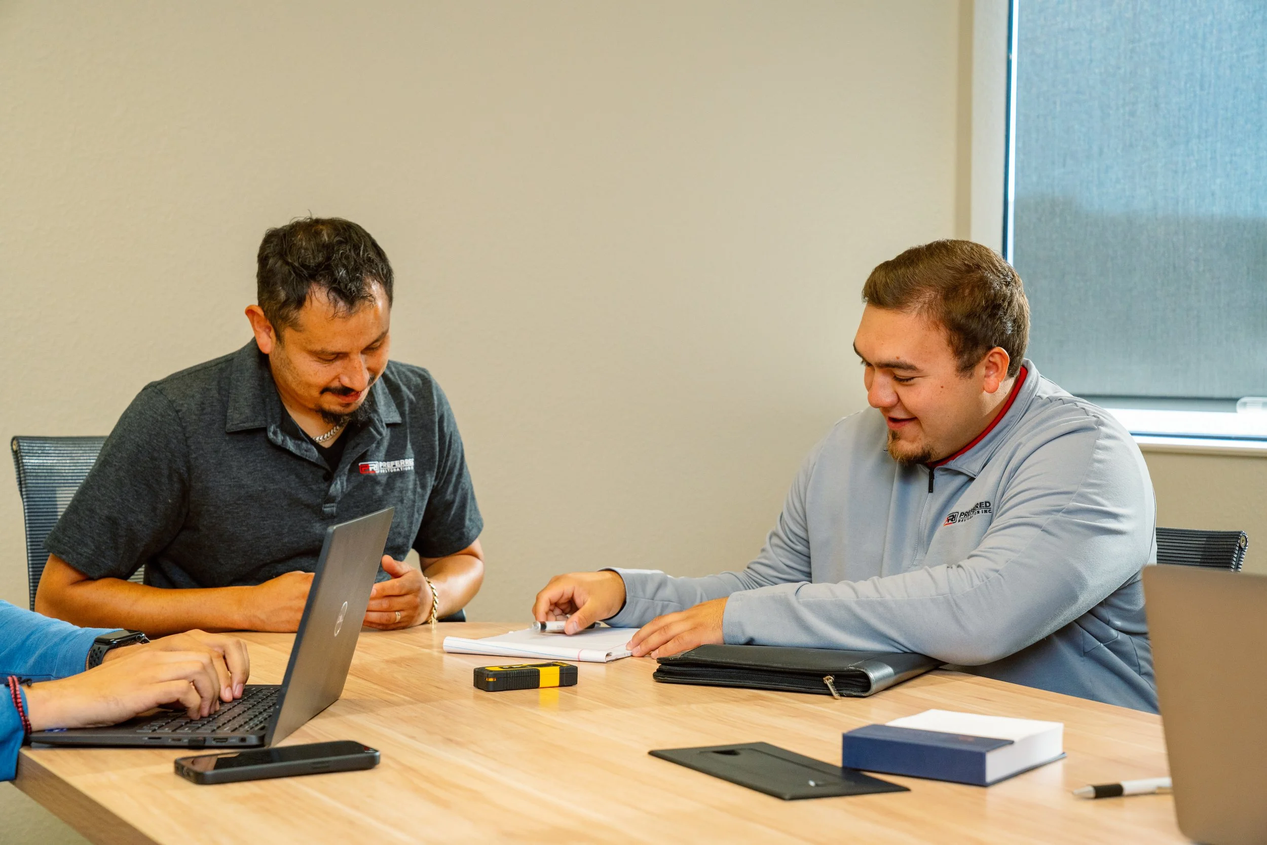 Two men sitting at a conference table, discussing documents, with laptops and notebooks in front of them.