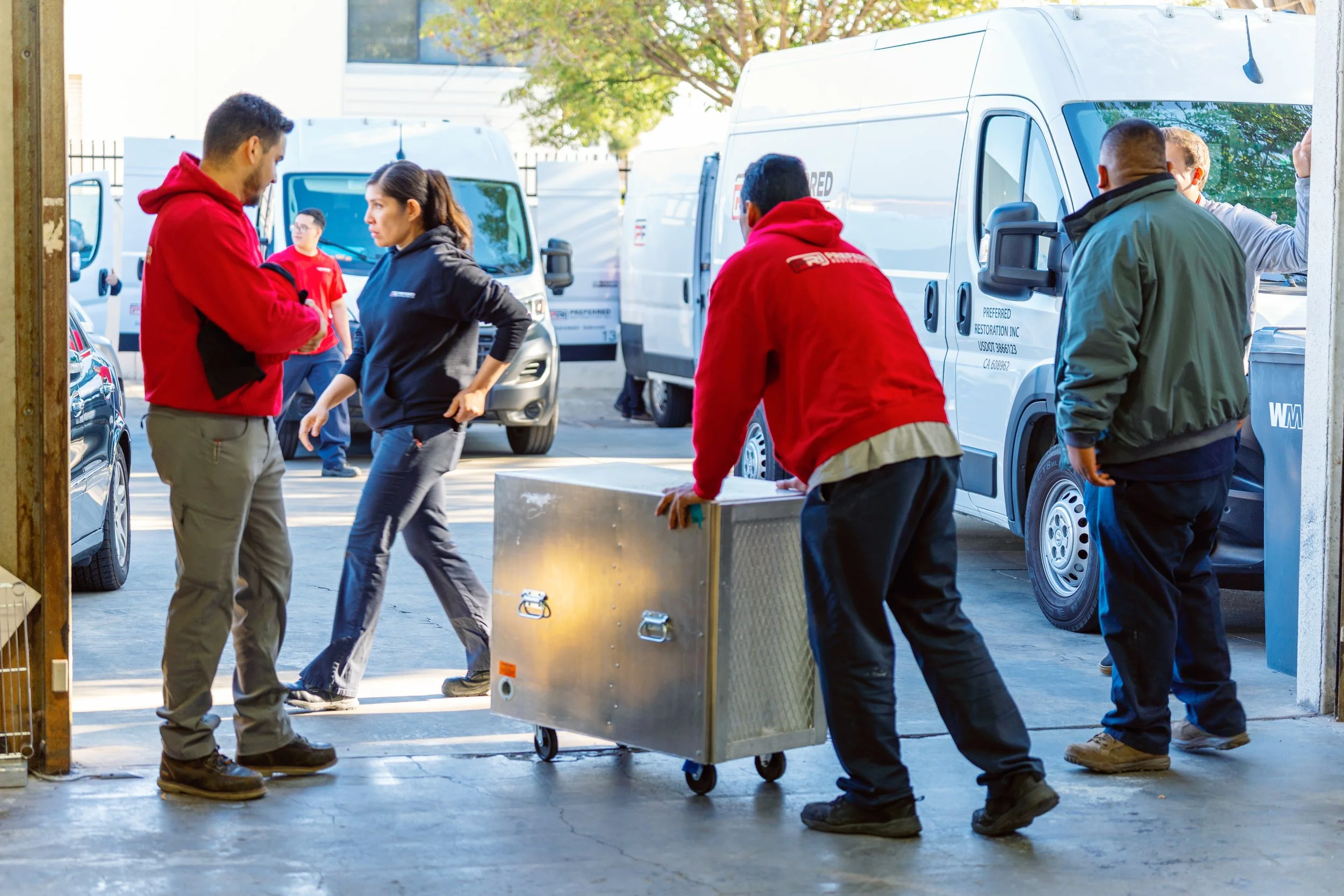 People loading a metal box on a dolly outside a warehouse, with service vans and other people in the background.
