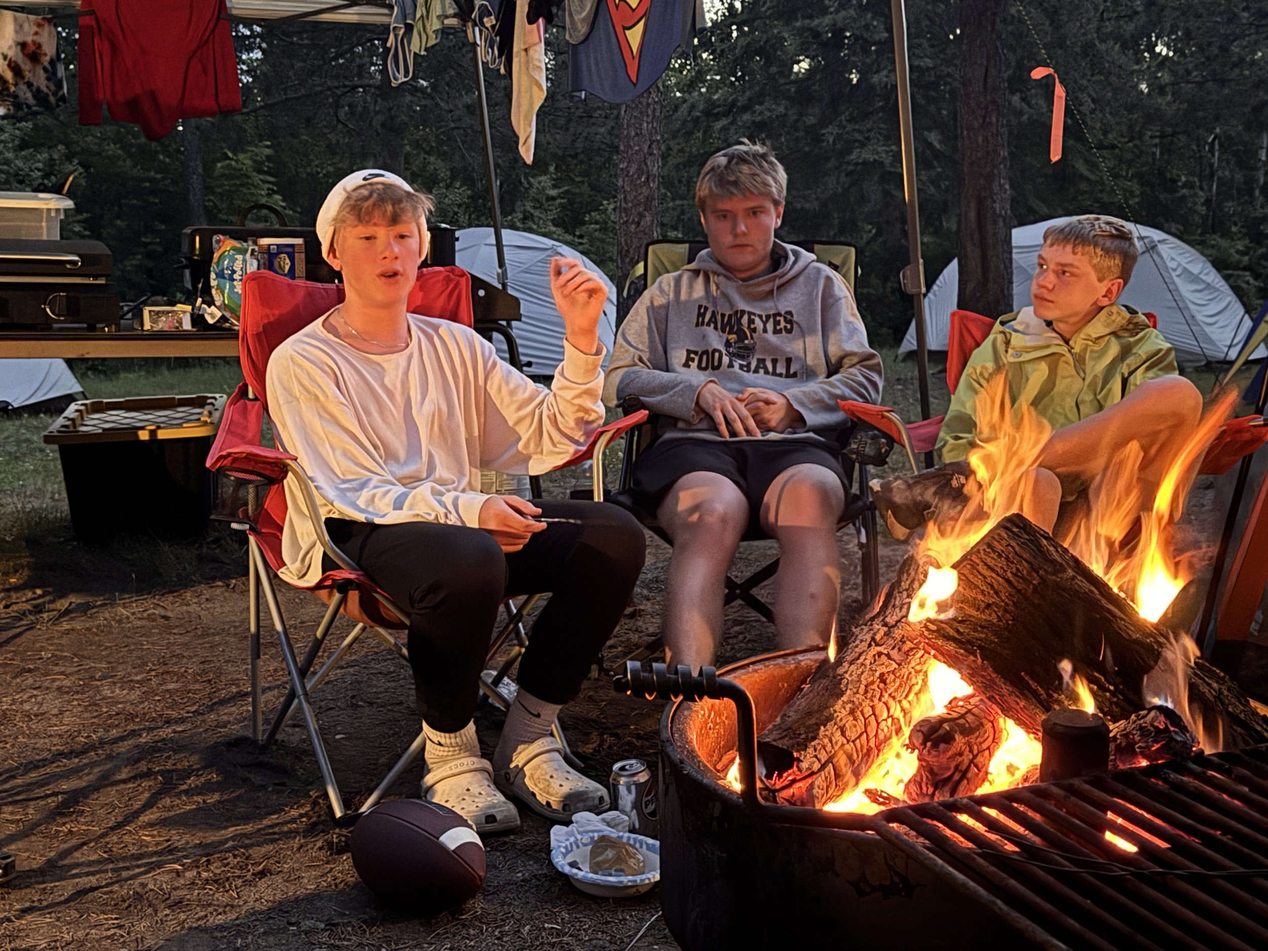 Three boys sitting around a campfire in a wooded campsite with tents in the background. One boy is speaking, wearing a white long-sleeve shirt and a beanie, while the other two boys listen attentively. Various camping supplies and a football are on the ground nearby.