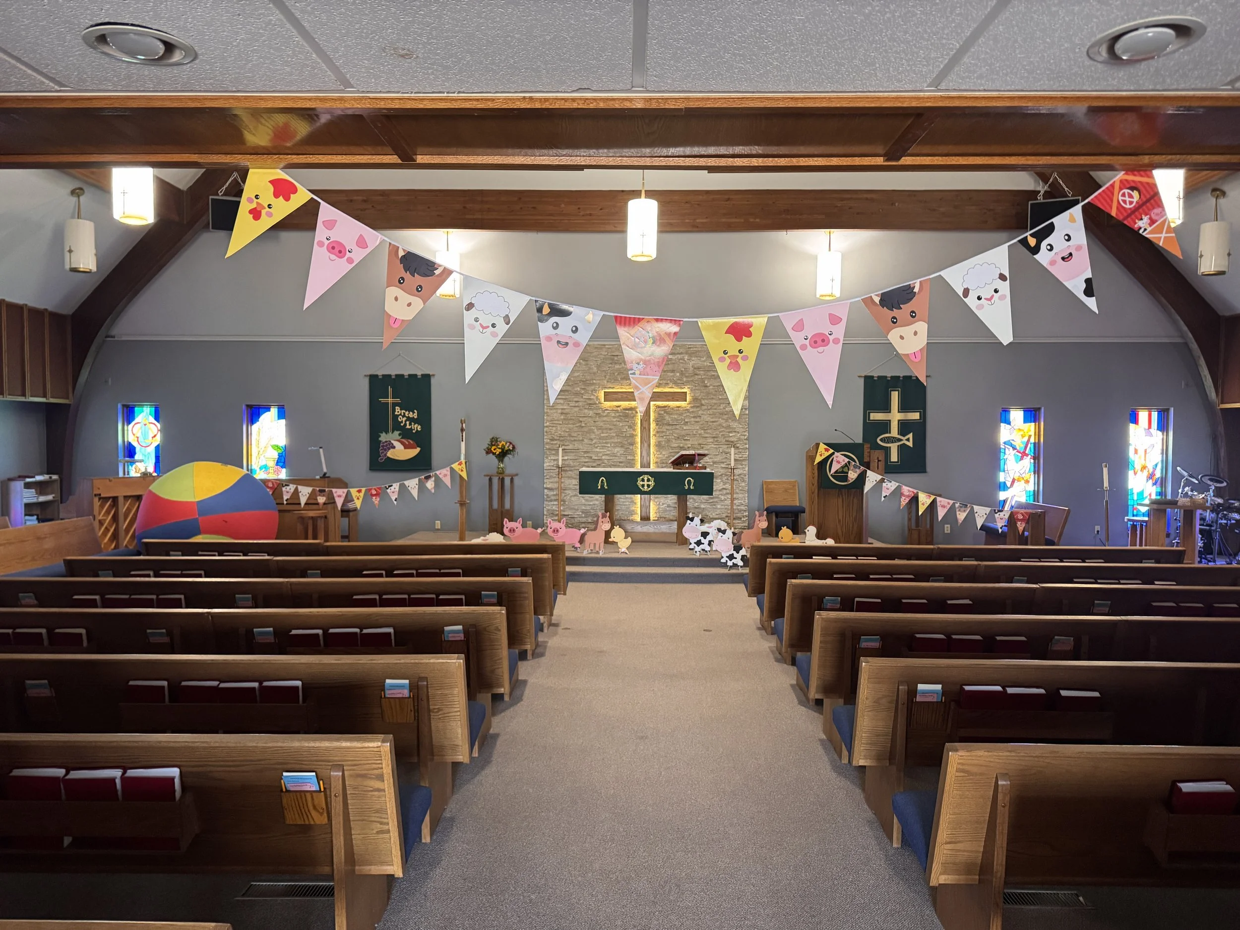 Inside a church with wooden pews, decorated with colorful pennant banners featuring farm animals and farm-themed illustrations, and stained glass windows.