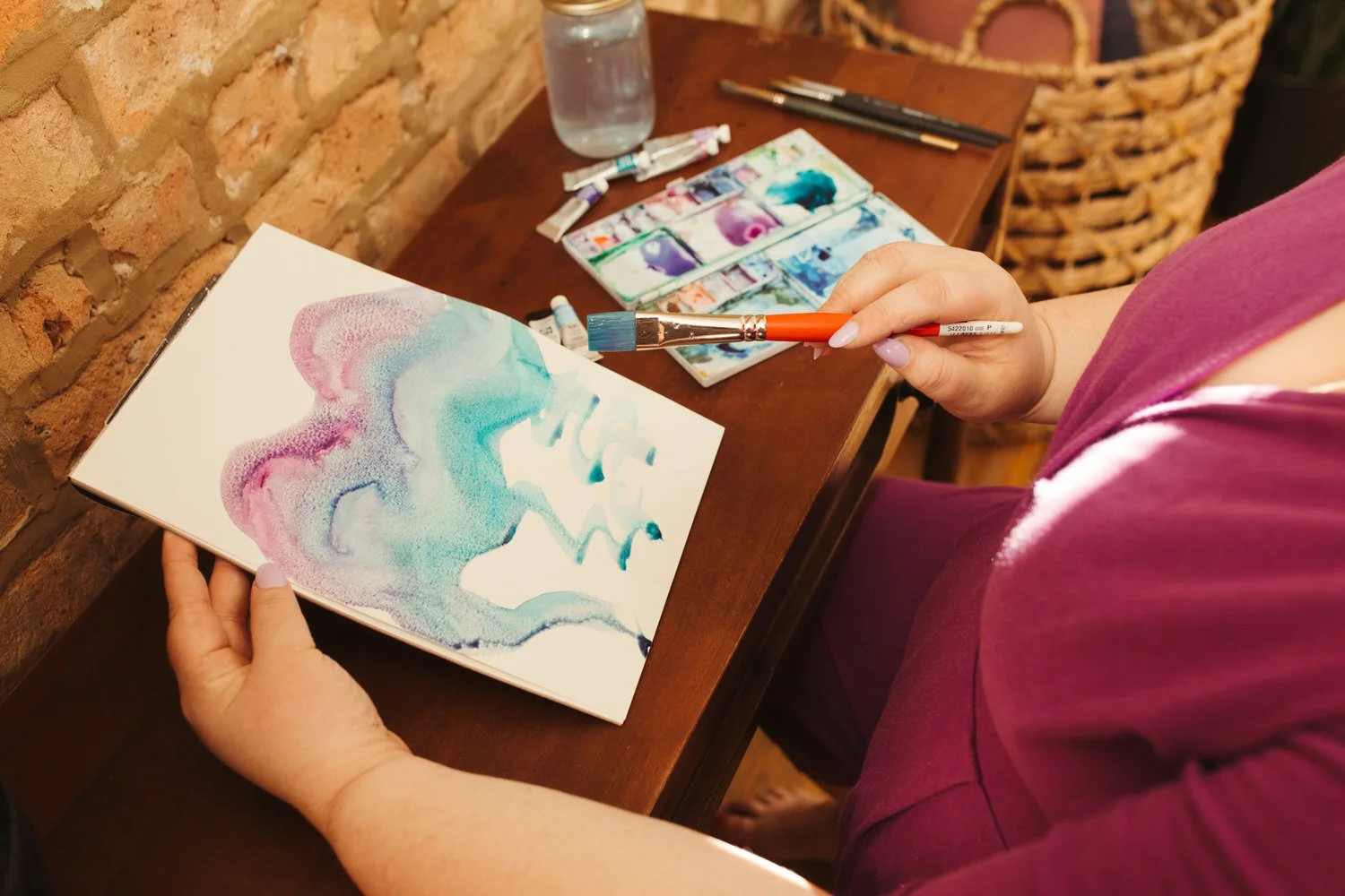 Person creating watercolor art on paper with paints and brushes on a wooden table.