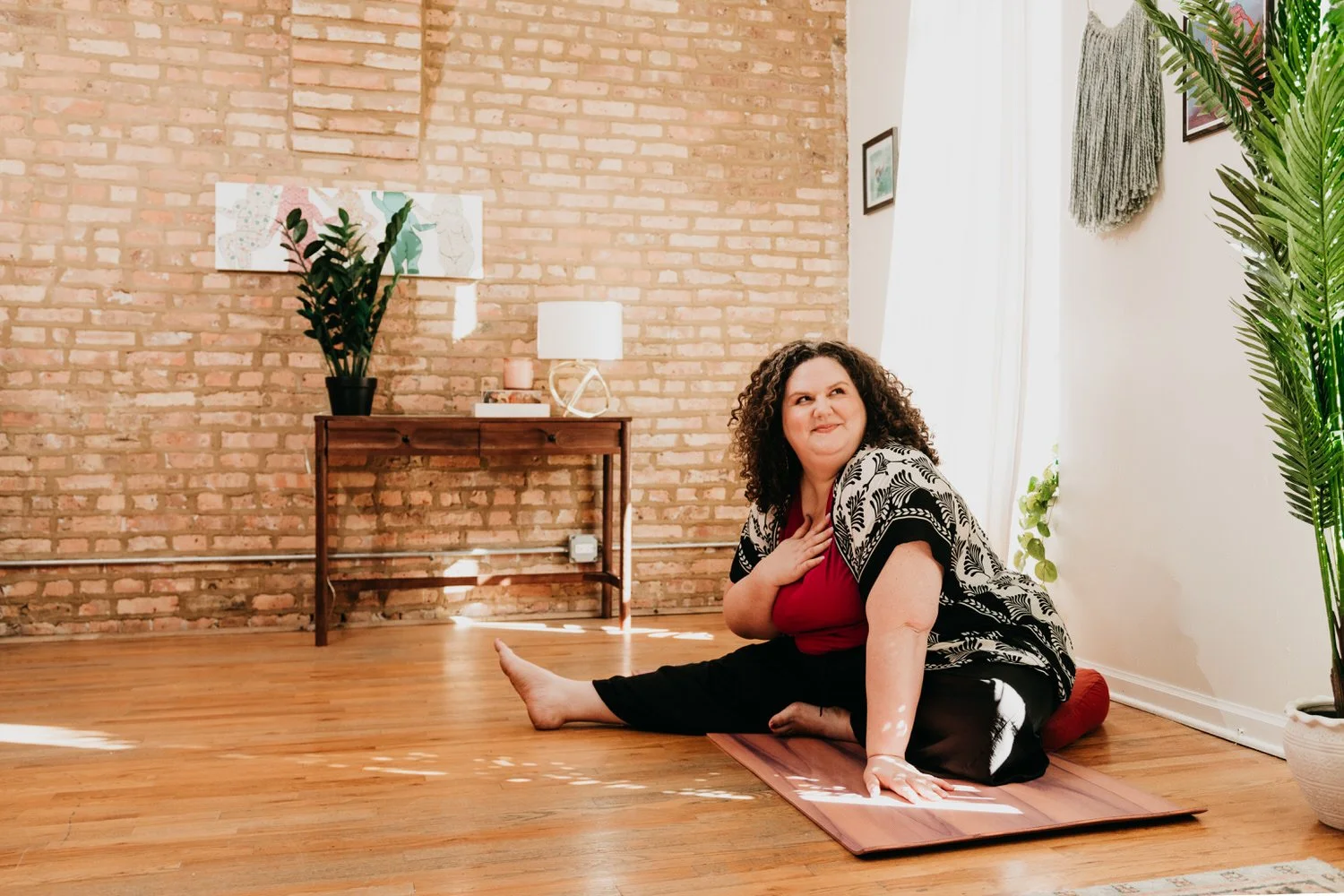 A woman with curly hair practicing yoga on a wooden floor mat in a room with exposed brick walls, a wooden table with a potted plant and a lamp, and large windows with white curtains, sunlight streaming in.