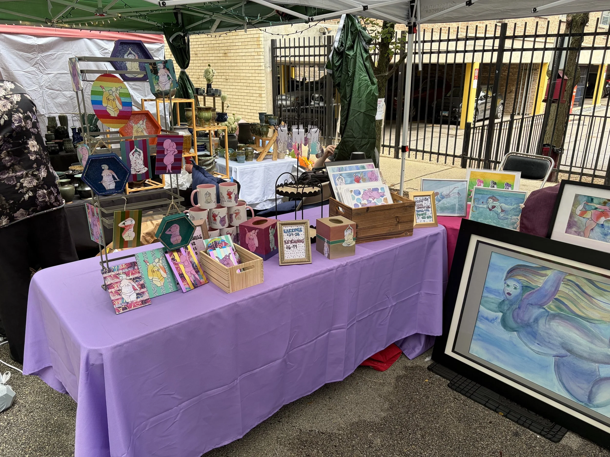 Naomi's colorful, joyful art is displayed in a booth, on a table with a purple table cloth. There are wood plaques, mugs, tissue box holders, prints, earrings, and keychains featuring fat subjects.