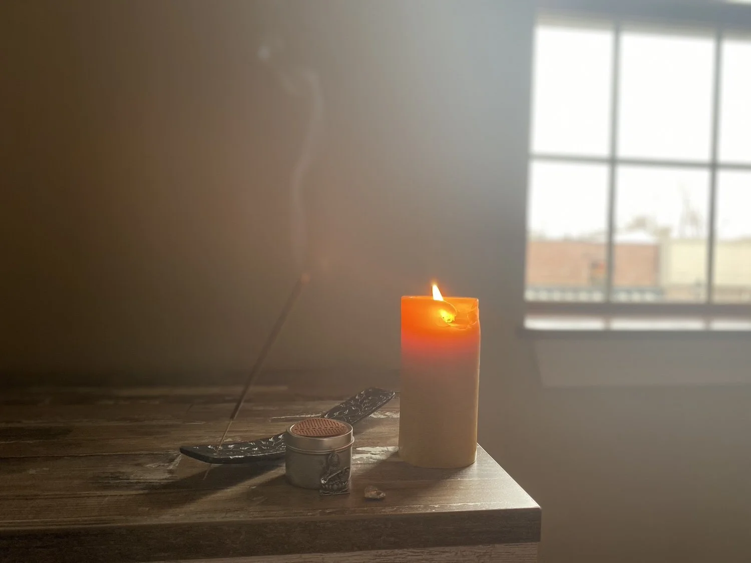 A lit orange candle on a wooden surface near a window, with an incense stick and a small container.