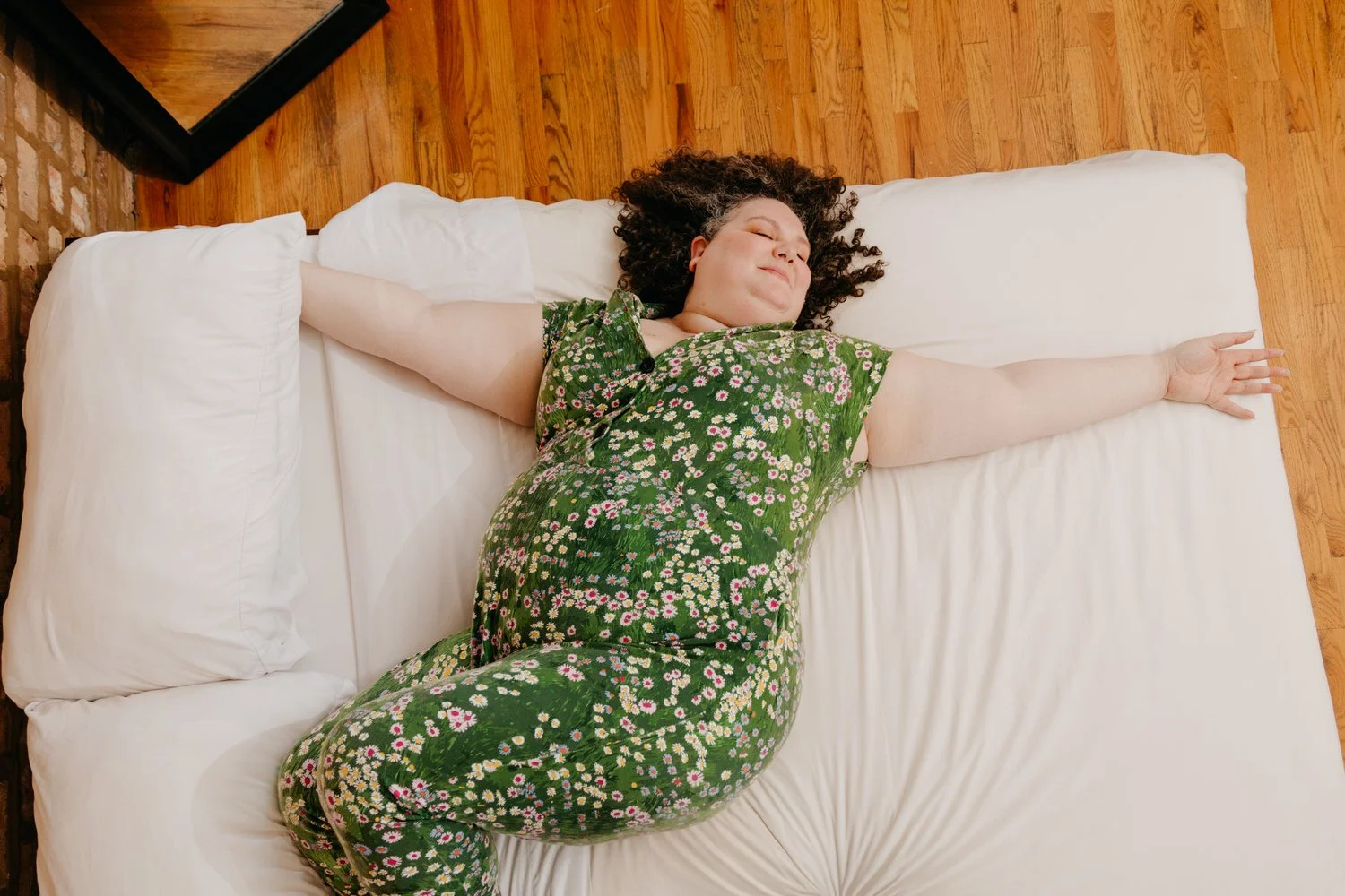 Woman with curly hair sleeping on her back in a floral green dress on a white bed with pillows and hardwood floor.
