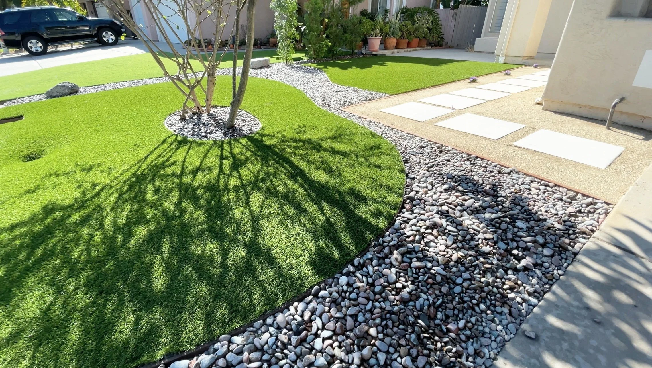A landscaped backyard with a small tree surrounded by white rocks, a green artificial lawn, and a gravel pathway with potted plants nearby.