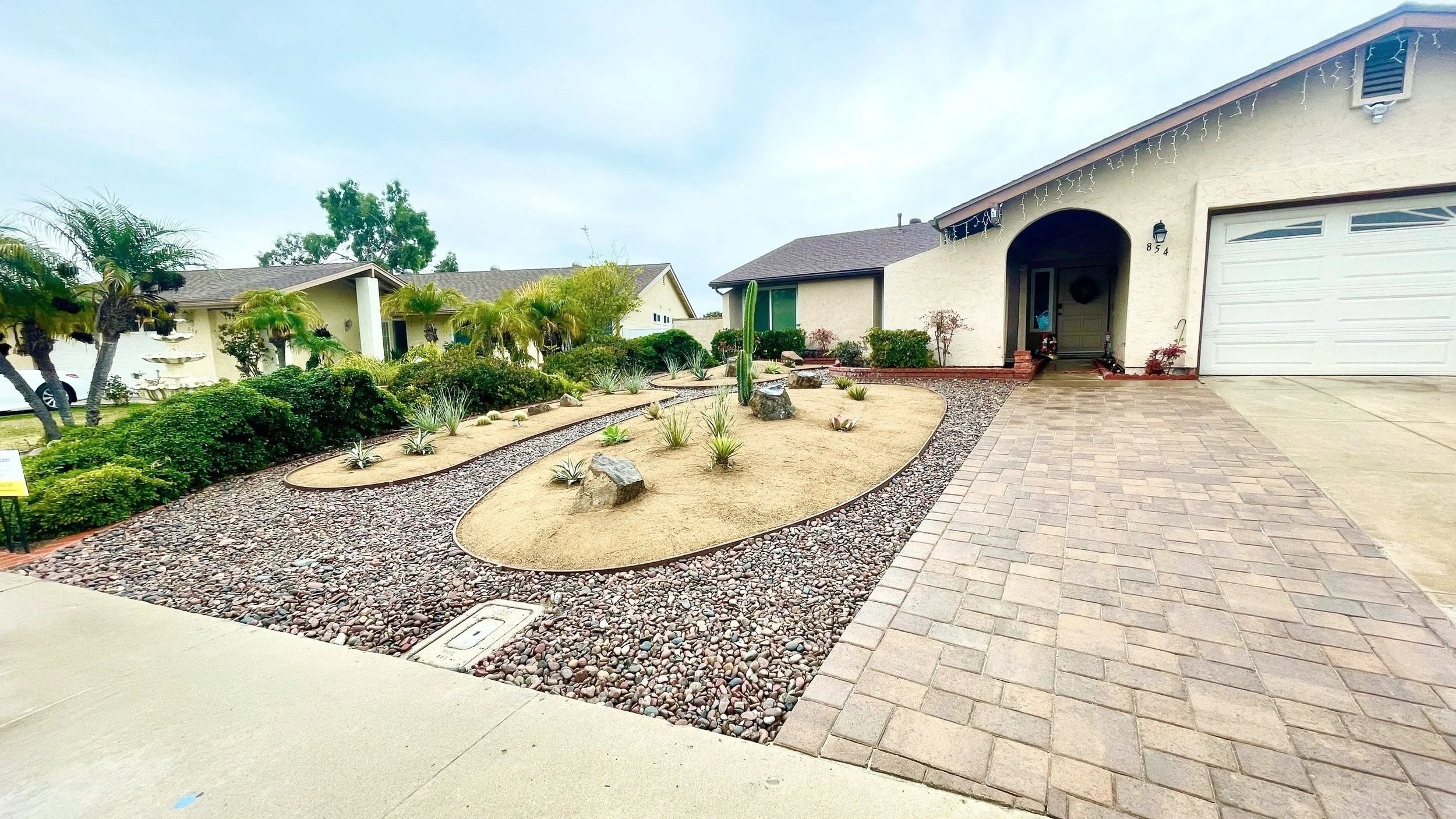 Front yard of a house with desert landscaping, including a cactus, rocks, and small plants, next to a brick driveway and walkway.