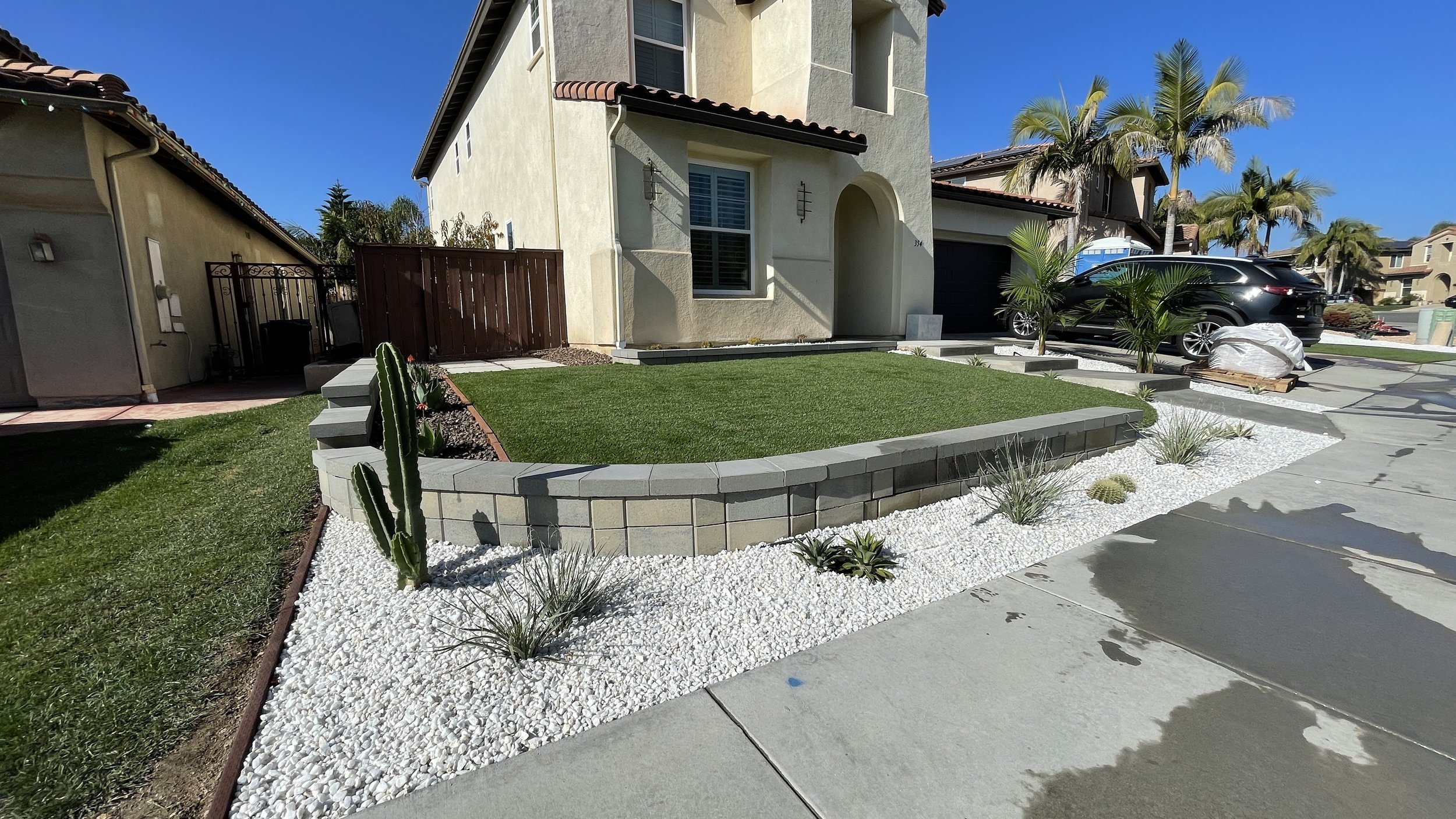 Front yard of a house with a small area of green grass, desert-style landscaping with white gravel, cacti, and small plants, a curved retaining wall made of gray bricks, and a concrete driveway with wet spots. There is a black car parked in the drive
