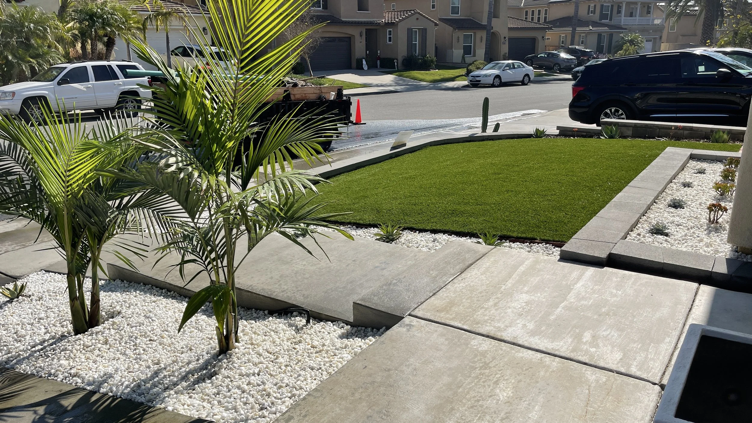 Front yard with green lawn, white gravel, and potted plants, with a sidewalk and street with parked cars in a suburban neighborhood.