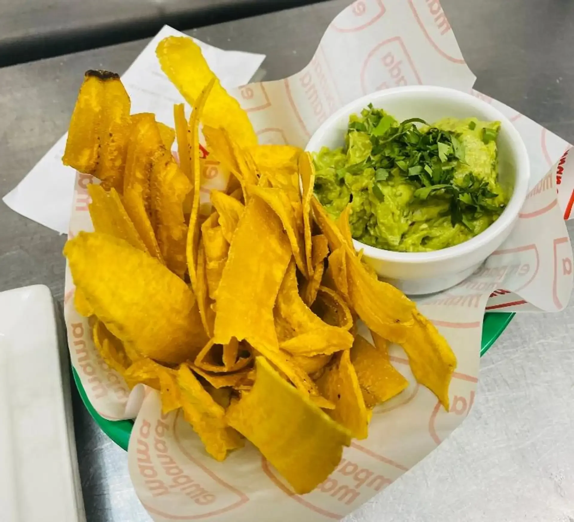 Crispy plantain chips with a side of fresh guacamole served in a branded paper basket at Empanada Mama Bergen Ave.