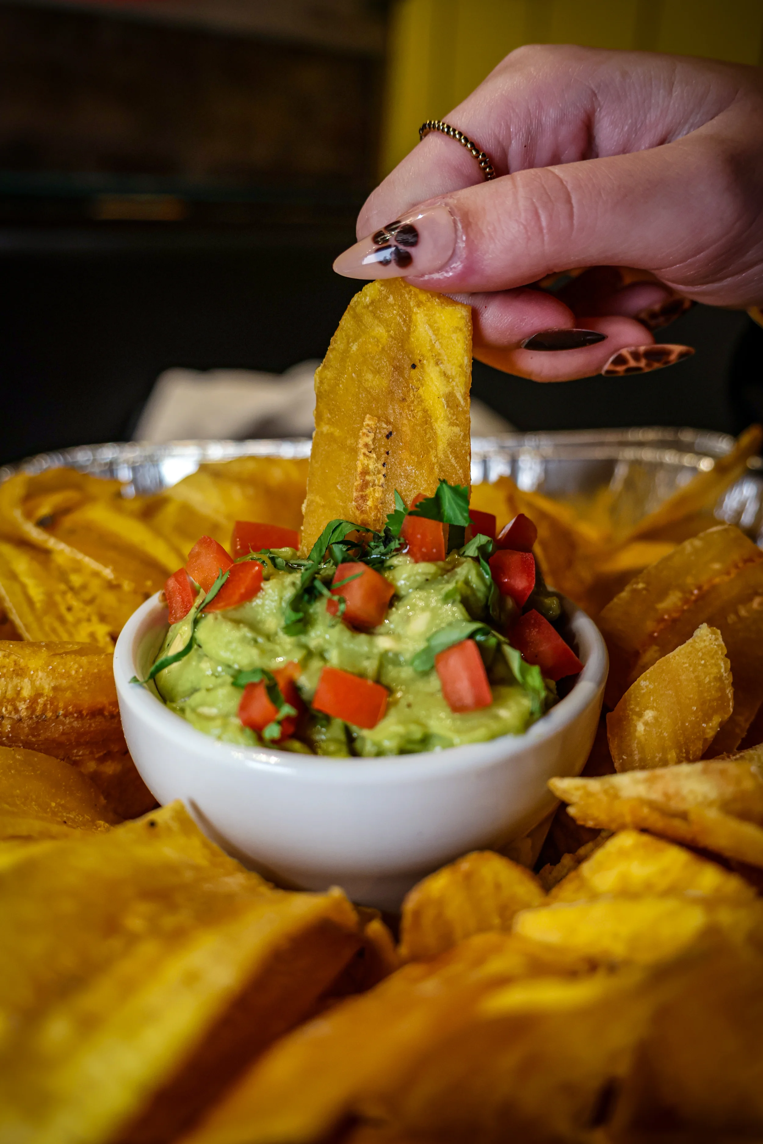 Empanada Mama fresh guacamole topped with diced tomatoes and cilantro, served with crispy plantain chips as a hand dips in for a bite.